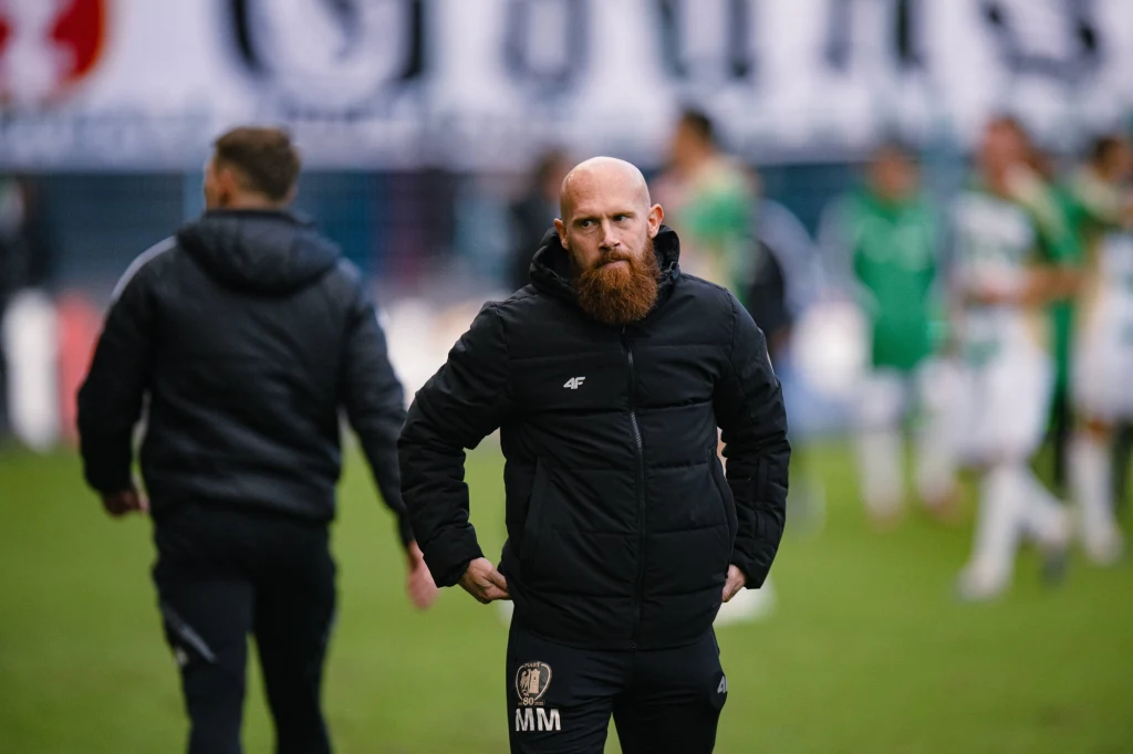 Max Moelder jako trener Piasta Gliwice wywalczył zaledwie jedno zwycięstwo w Ekstraklasie Łysy mężczyzna z rudą brodą w czarnej kurtce sportowej stoi na murawie stadionu, w tle kilku innych uczestników wydarzenia sportowego oraz rozmyty baner i publiczność.