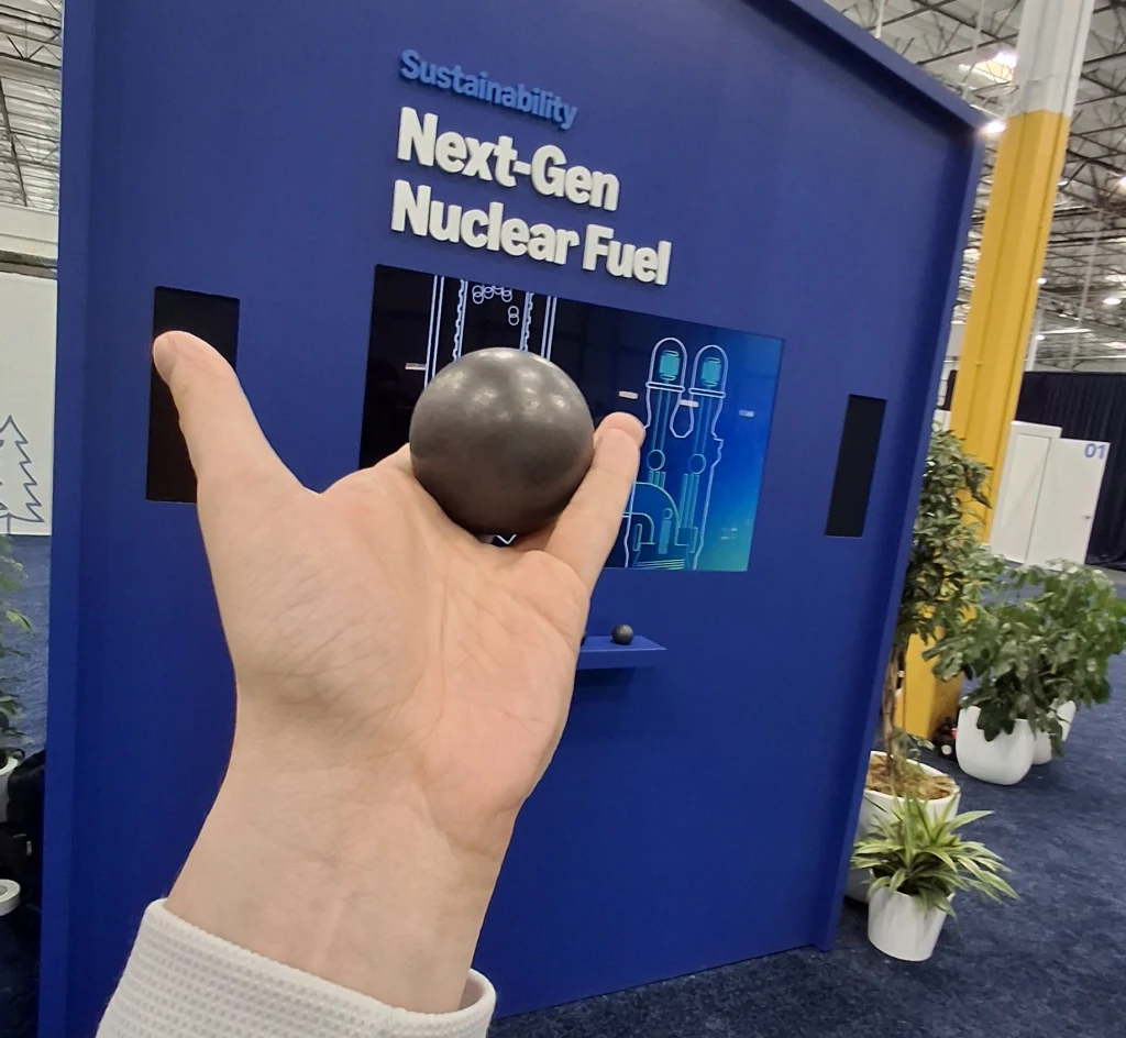 Ręka trzyma czarną kulę na tle niebieskiego stoiska z napisem Next-Gen Nuclear Fuel; w tle znajdują się rośliny doniczkowe, plansza z grafiką technologiczną oraz część hali wystawienniczej.
