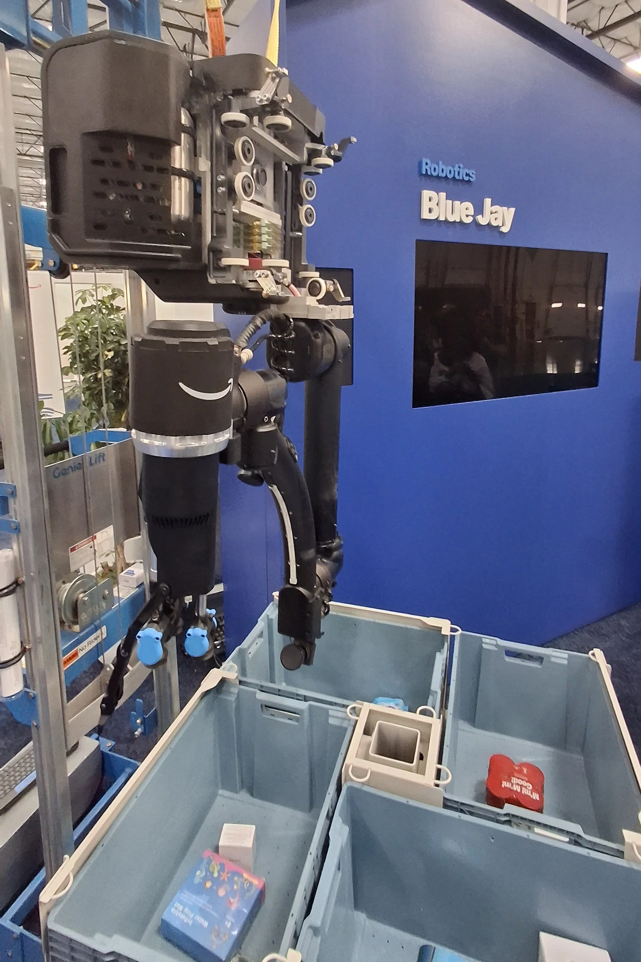 Nowoczesny robot przemysłowy wyposażony w chwytaki sortuje przedmioty w pojemnikach na tle niebieskiego stoiska wystawowego z napisem Blue Jay.