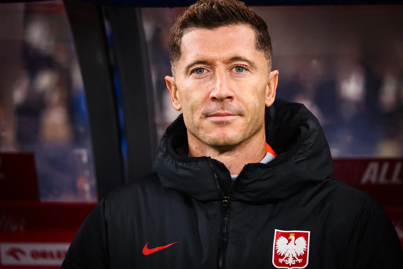 Mężczyzna w kurtce sportowej z godłem Polski na piersi, patrzący prosto w obiektyw, w tle rozmyta przestrzeń stadionowa.