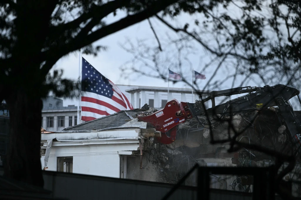 Prace rozbiórkowe wschodniego skrzydła Białego Domu w Waszyngtonie ruszyły na początku tygodnia Maszyna wyburzająca burzy fragment budynku z amerykańską flagą na pierwszym planie, w tle widoczny jest biały gmach oraz powiewające flagi USA.