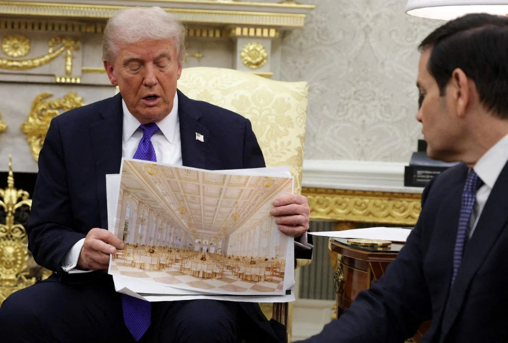 Dwóch mężczyzn siedzi w elegancko urządzonym pomieszczeniu. Jeden z nich trzyma i prezentuje arkusz papieru z wydrukowanym obrazem dużej, jasnej i ozdobnej sali, przeznaczonej do organizacji przyjęć lub wydarzeń. W tle widoczny luksusowy wystrój wnętrz...