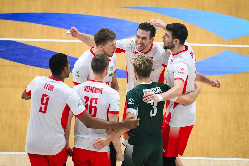 Grupa polskich siatkarzy ubranych w biało-czerwone stroje cieszy się razem na boisku, otaczając się ramionami po zdobytym punkcie, na drewnianym parkiecie widoczne są niebieskie elementy graficzne.