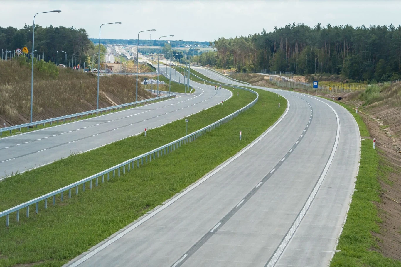 Ogłoszono ważny przetarg w sprawie realizacji odcinka drogi ekspresowej S11./zdjęcie ilustracyjne/ Dwupasmowa, pusta autostrada prowadząca przez zieloną okolicę, otoczona barierkami i oświetleniem ulicznym, z lasem i otwartą przestrzenią w tle.