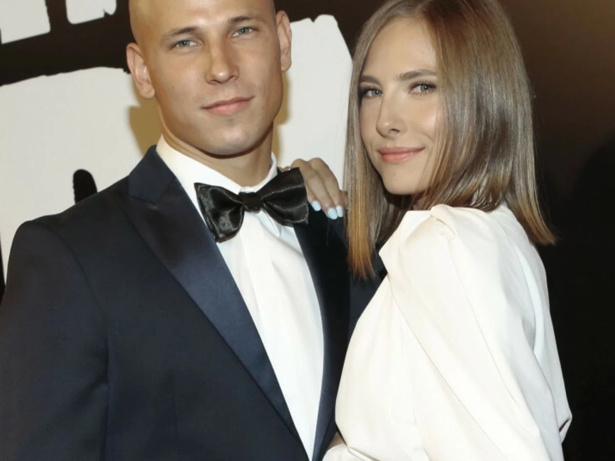 Józef Pawłowski i Klaudia Szafrańska Para elegancko ubranych osób pozuje na tle czarno-białej grafiki, mężczyzna ma na sobie smoking z muszką, kobieta ubrana jest w białą suknię z długim rękawem, oboje uśmiechnięci i przytuleni do siebie.