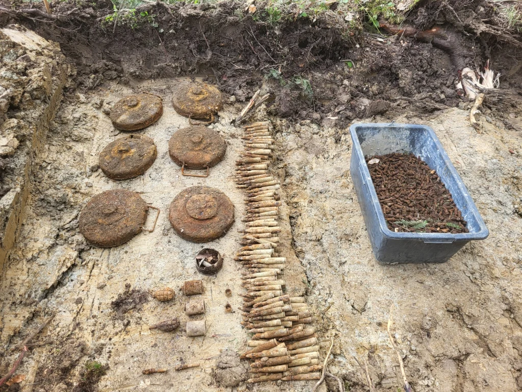 Rząd min przeciwpiechotnych, kilkadziesiąt pocisków oraz pojemnik wypełniony skorodowanymi łuskami i nabojami leżą na odkrytej ziemi, prawdopodobnie pochodzą ze znaleziska z okresu wojny.