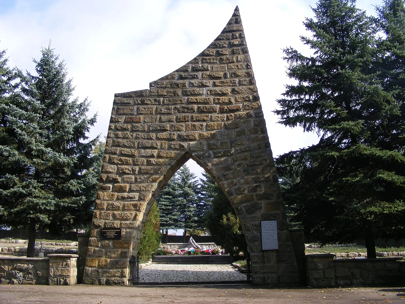 Masynistyczny kamienny łuk prowadzący na cmentarz wojskowy, otoczony wysokimi świerkami, w oddali widoczny pomnik i uporządkowane groby z kwiatami.