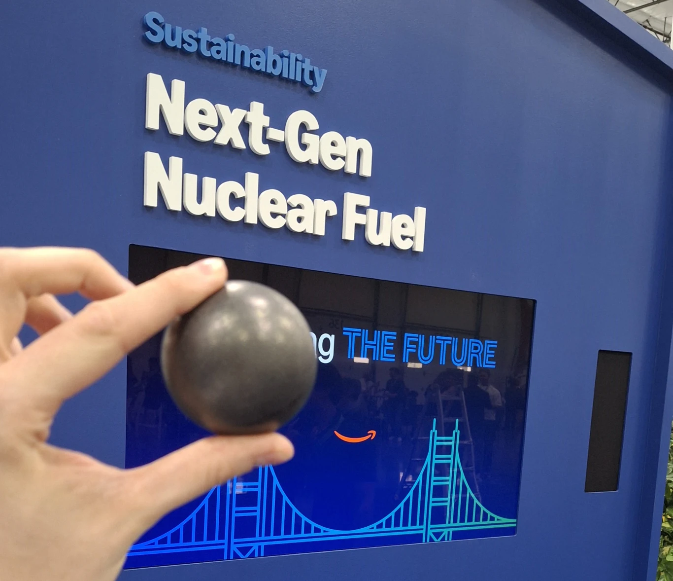Ta jedna kulka może być przyszłością energii nuklearnej. Oto co zaprezentowano na Amazon Delivering the Future Ręka trzymająca kulę w ciemnym kolorze na tle ściany z napisem dotyczącym zrównoważonego rozwoju oraz nowoczesnego paliwa jądrowego, podświetlona grafika mostu i hasło o przyszłości.