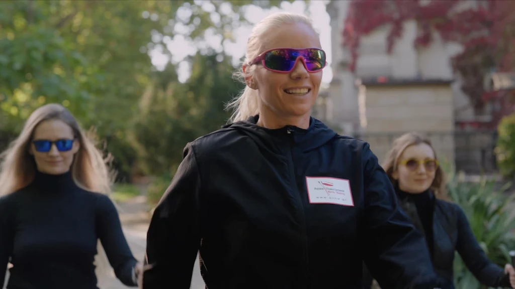 Nordic walking to pełnoprawna aktywność sportowa, angażująca aż 90 proc. mięśni Trzy kobiety idące alejką w parku, ubrane w sportowe stroje i okulary przeciwsłoneczne, główna postać uśmiecha się, w tle widoczna zieleń oraz budynek pokryty fragmentarycznie czerwoną winoroślą.