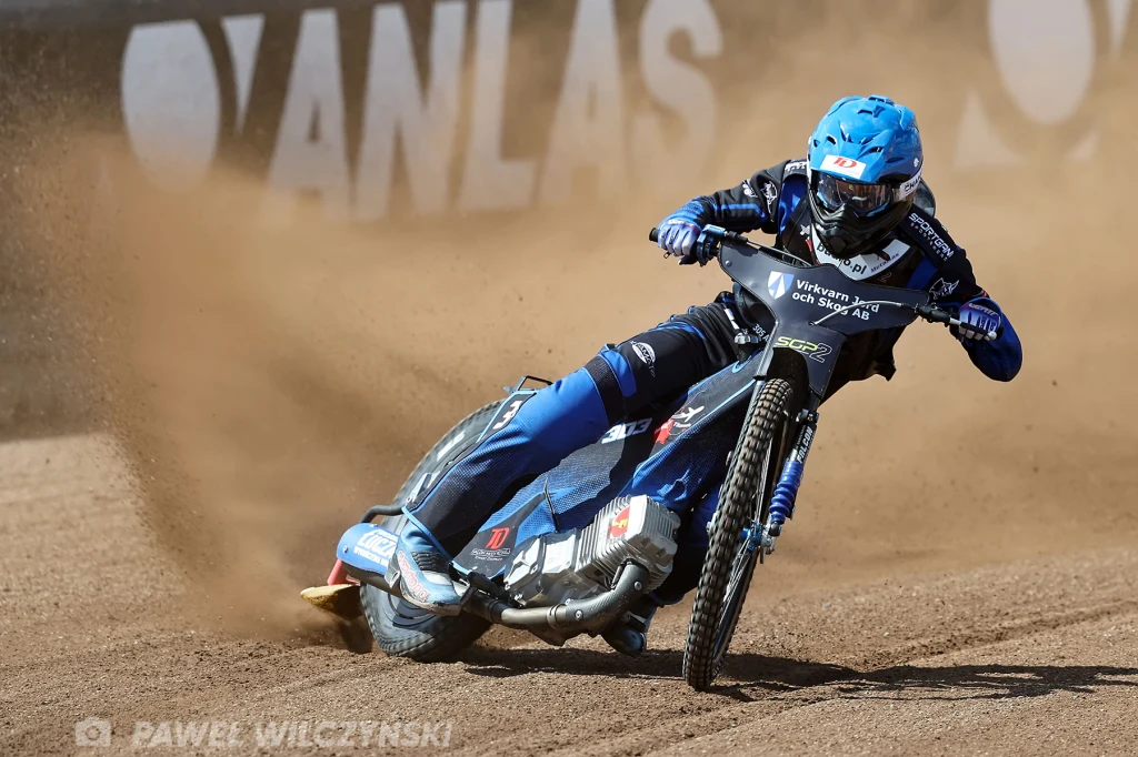 Antoni Mencel w SGP2. Motocyklista w niebieskim stroju pokonuje zakręt na torze żużlowym, wzbijając kurz spod tylnego koła, w tle widać fragment reklamowego baneru.