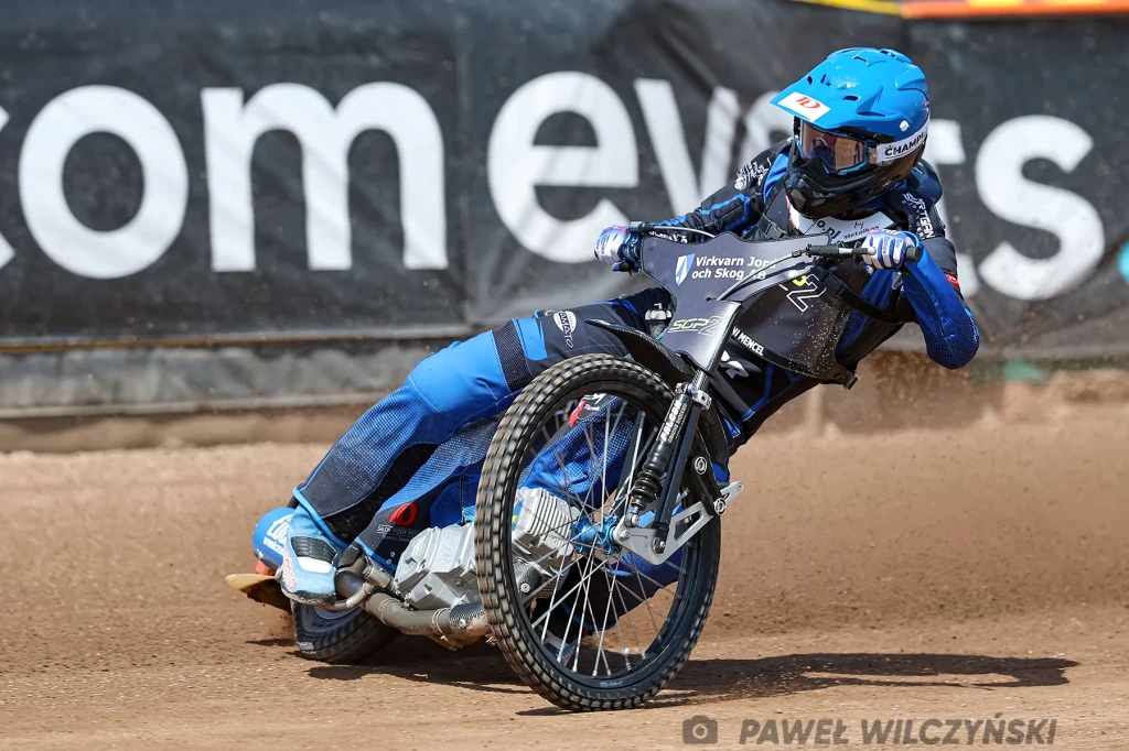 Antoni Mencel w SGP2. Zawodnik żużlowy w pełnym ekwipunku podczas dynamicznego zakrętu na torze, motocykl przechylony blisko nawierzchni, tło stanowi rozmyty fragment reklamy i bandy torowej.