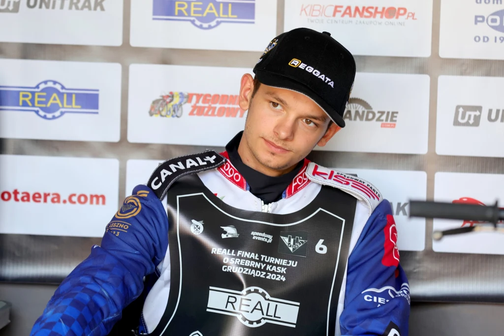 Antoni Mencel. Zawodnik żużlowy w stroju sportowym, siedzący na ławce, w tle tablica z logotypami sponsorów i partnerów wydarzenia sportowego.