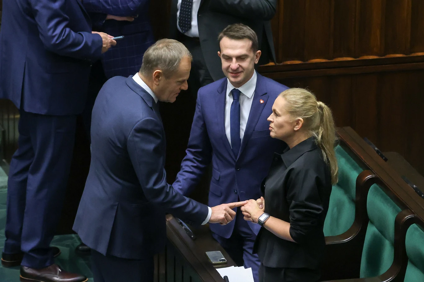 Donald Tusk, Barbara Nowacka oraz Adam Szłapaka Trójka polityków w eleganckich garniturach i koszuli prowadzi rozmowę w ławach parlamentu, skupiając na sobie uwagę, a reszta osób widoczna jest w tle.