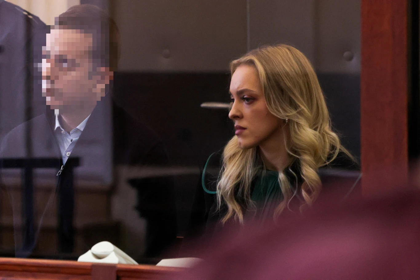 Sebastian M. wraz ze swoją adwokat Katarzyną Hebdą na sali sądowej Kobieta o blond włosach siedzi przy stole za szybą w pomieszczeniu sądowym, obok niej znajduje się mężczyzna z zamazaną twarzą. Kobieta jest skupiona i patrzy przed siebie, cała scena jest formalna i urzędowa.