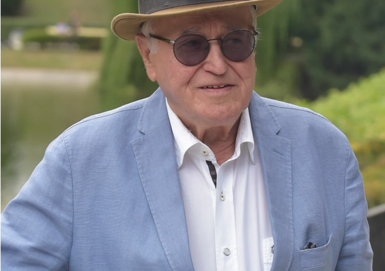 Jan Nowicki Starszy mężczyzna w okularach przeciwsłonecznych oraz jasnoniebieskiej marynarce, ubrany elegancko, z kapeluszem na głowie. W tle widoczna zielona roślinność i lekko rozmyty krajobraz parkowy.