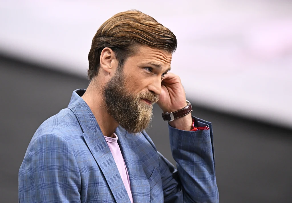 Andrzej Wrona Brodaty mężczyzna w niebieskiej marynarce, jasnoróżowym T-shircie i zegarku na ręku, lekko przechylony, z ręką przy uchu, o zamyślonym lub zaniepokojonym wyrazie twarzy, na rozmytym tle.