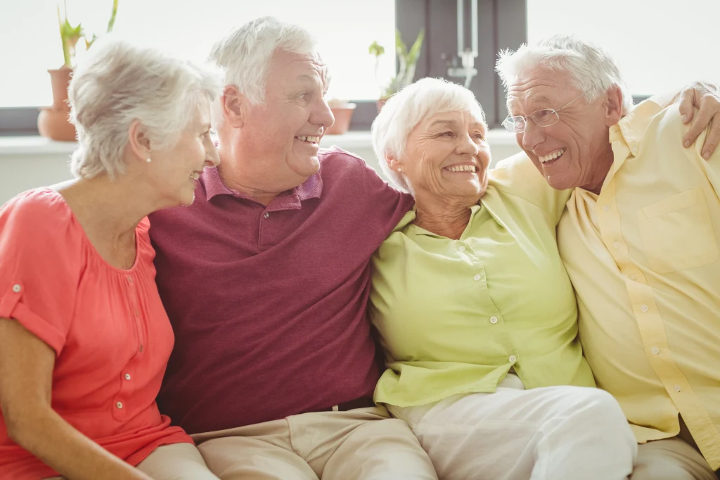 Czwórka starszych, uśmiechniętych osób siedzących razem na kanapie, ubranych w kolorowe ubrania, przytulających się i prowadzących radosną rozmowę, w tle widać kwiaty doniczkowe na parapecie.