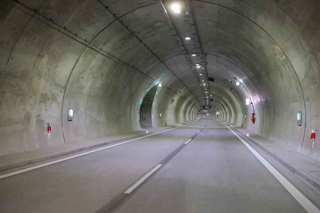 Tunel na obejściu Węgierskiej Górki w masywie Baraniej Góry Betonowy tunel drogowy z oświetleniem sufitowym, pusty, z widoczną białą linią oddzielającą pasy oraz zamontowanymi urządzeniami bezpieczeństwa i gaśniczymi na ścianach.