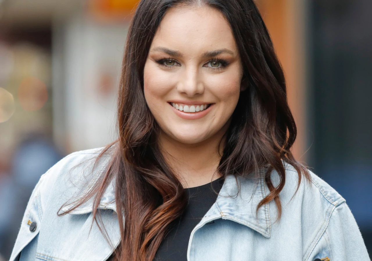 Ewa Farna Młoda kobieta o długich, ciemnych włosach i delikatnym makijażu, uśmiechnięta, ubrana w jasną kurtkę jeansową, stoi na tle rozmytego miejskiego krajobrazu.