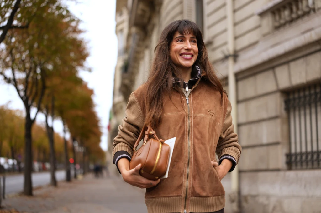 Kobieta z długimi, ciemnobrązowymi włosami uśmiecha się spacerując miejską ulicą, ubrana w brązową kurtkę, z torebką w dłoni oraz liśćmi drzew po bokach chodnika.