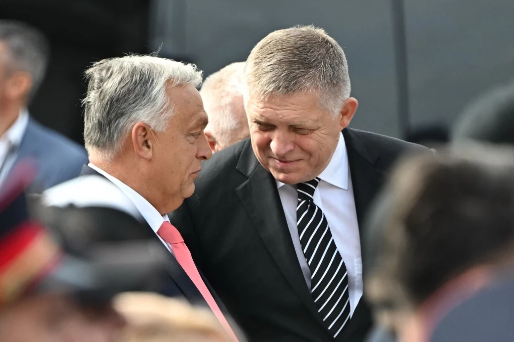 Premierzy Węgier i Słowacji: Viktor Orban i Robert Fico Dwóch mężczyzn w garniturach prowadzi rozmowę, stojąc blisko siebie, jeden z nich ma siwe włosy i nosi jasnoczerwoną krawatę, drugi ma krótkie blond włosy i pasiastą czarno-białą krawatę, w tle widoczne inne osoby i zamazane elementy otoczenia.
