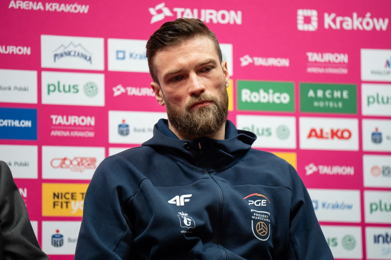 Andrzej Wrona w barwach PGE Projektu Warszawa, w którym zakończył karierę Mężczyzna z brodą ubrany w sportową bluzę z logo klubu PGE siedzi na tle różowej ścianki z logotypami sponsorów podczas konferencji prasowej.