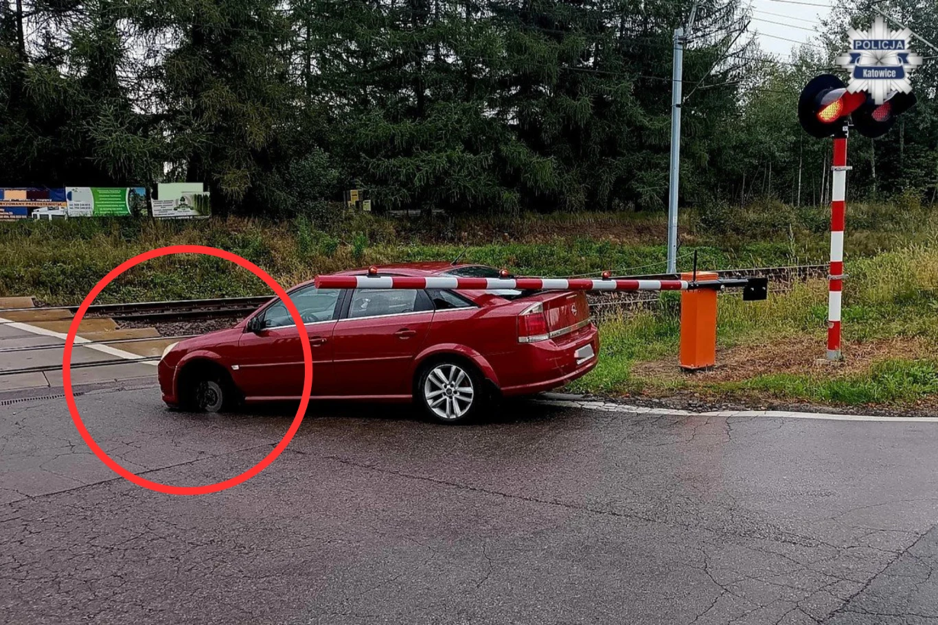Koło odpadło z samochodu na środku przejazdu kolejowego. Auto szczęśliwie udało się zepchnąć przed nadjeżdżającym pociągiem. Czerwony samochód osobowy stoi na zamkniętym przejeździe kolejowym, przód auta znajduje się pod opuszczonym szlabanem, a przednie lewe koło pojazdu wpadło w szczelinę torowiska. Widoczne są znaki kolejowe i sygnalizacja, a dalszy plan zajmuje zieleń or...