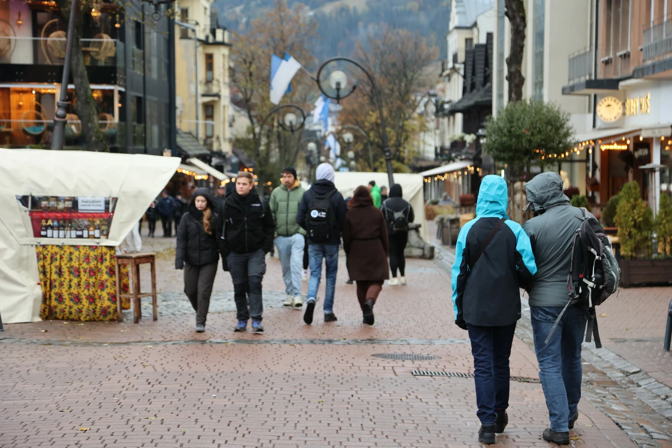 Mimo jesieni w Zakopanem turystów nie brakuje. Ulica miasta z przechodniami ubranymi w ciepłe kurtki, kilka stoisk ulicznych po lewej stronie, budynki po obu stronach ulicy, nastrojowe lampy i jesienna aura w tle.