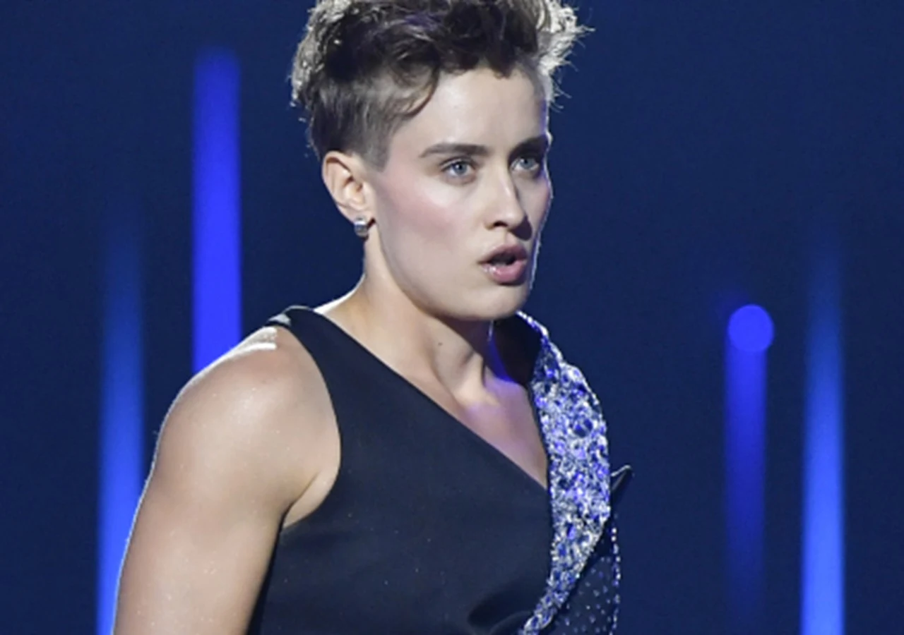 Katarzyna Zillmann Osoba z krótkimi włosami w czarnej stylizacji z błyszczącym elementem na ramieniu stoi na scenie w mocnym świetle z niebieskimi światłami w tle, wyrażając skupienie i emocje.