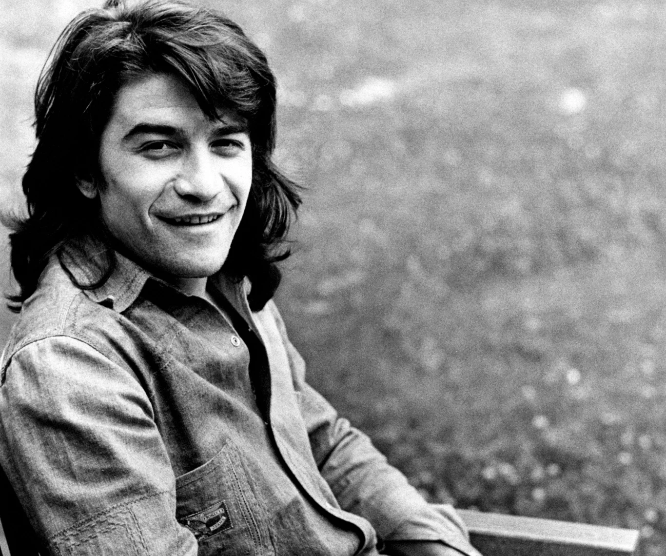 Drupi w 1974 r. Młody mężczyzna z długimi ciemnymi włosami siedzący na ławce, uśmiechnięty, ubrany w koszulę, w tle rozmyta naturalna sceneria.