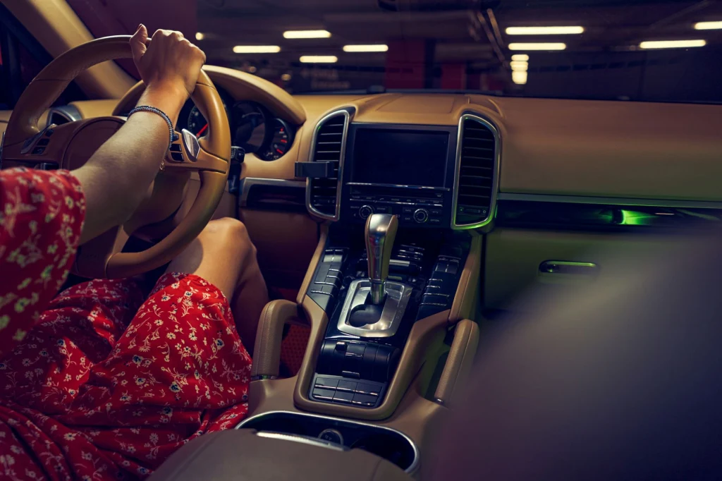 System awaryjnego hamowania. Na czym polega? Wnętrze luksusowego samochodu z jasnobrązową tapicerką, kobieta w czerwonej sukience w kwiaty siedzi za kierownicą, trzymając ją jedną ręką, deska rozdzielcza wykończona eleganckimi materiałami.