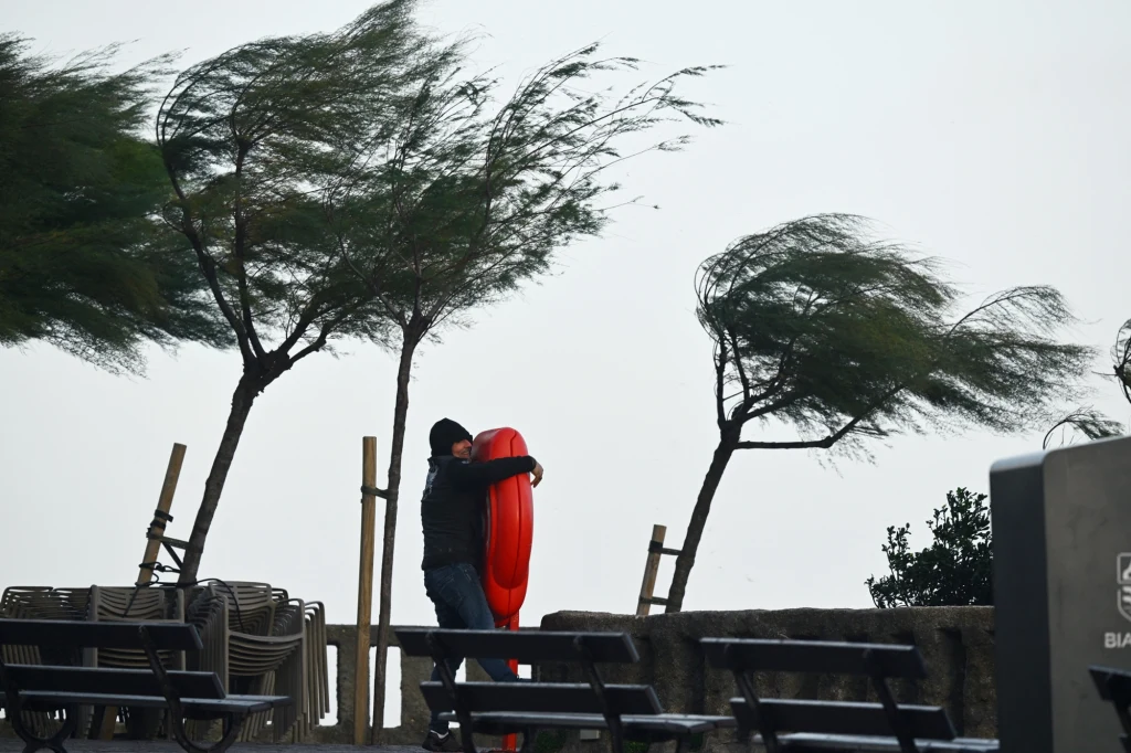 Mężczyzna trzymający czerwoną boję ratunkową na tle mocno wiejącego wiatru, drzewa wygięte pod naporem wiatru, puste ławki i ponure niebo.
