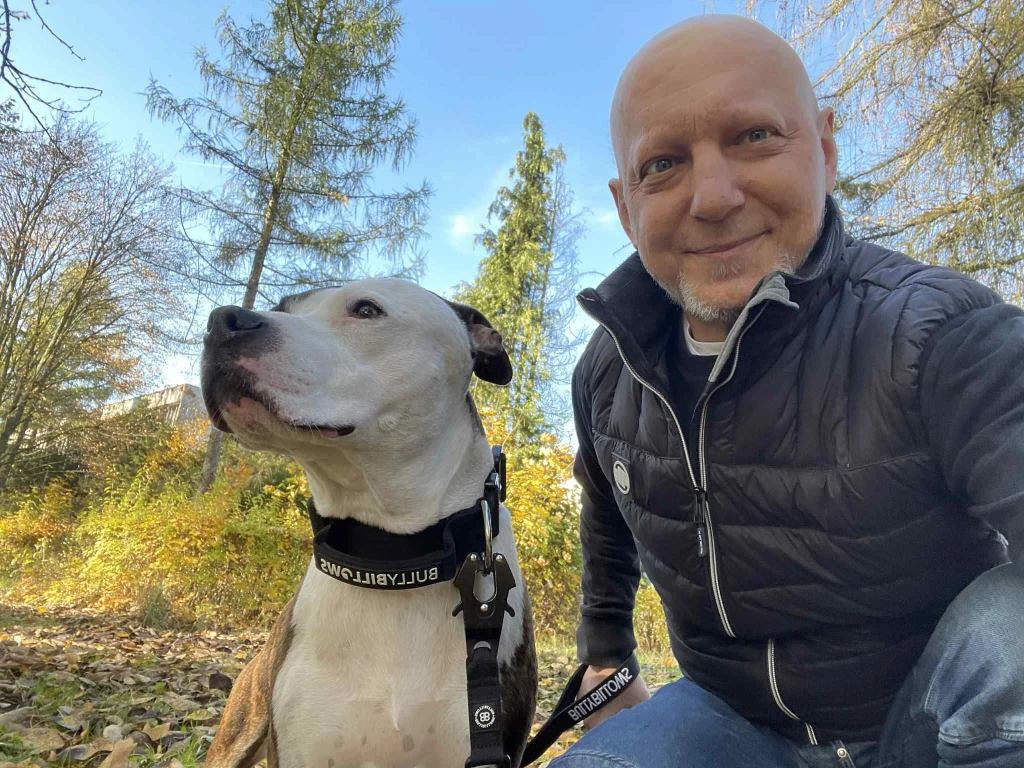 Uśmiechnięty mężczyzna siedzi obok psa rasy pitbull na tle jesiennych drzew, oboje wyglądają spokojnie i zrelaksowani, a dookoła leżą opadłe liście.