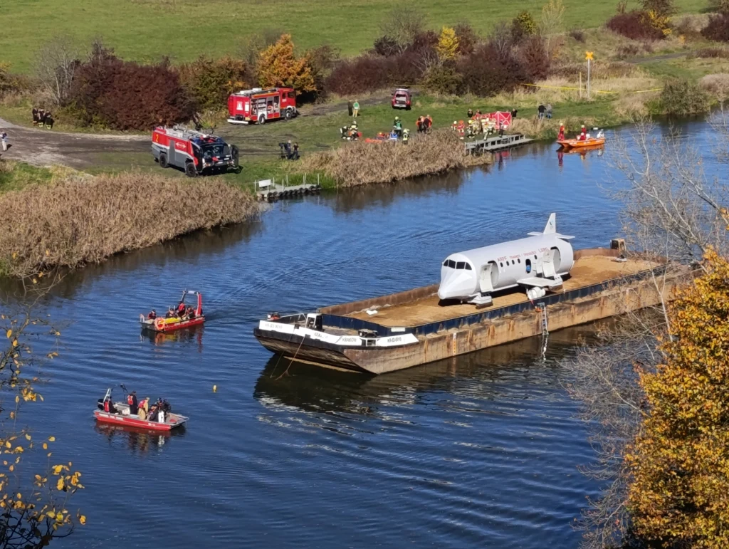Wrak samolotu przewożony barką po rzece, otoczony jednostkami ratunkowymi na wodzie oraz pojazdami straży pożarnej na brzegu. W tle widoczni są ratownicy i jesienna roślinność.