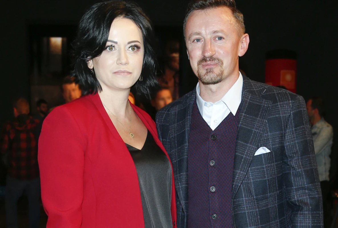 Kobieta z krótkimi ciemnymi włosami w czerwonej marynarce i mężczyzna w granatowej marynarce w kratę stoją razem na tle ciemnego wnętrza, oboje patrzą w kierunku obiektywu.