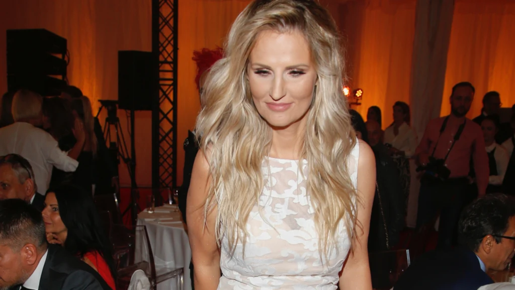 Dominika Tajner Kobieta z długimi blond włosami ubrana w jasną, elegancką suknię stoi w jasnym wnętrzu podczas formalnej imprezy, w tle widoczni są inni goście siedzący przy stołach oraz elementy dekoracji świetlnej.