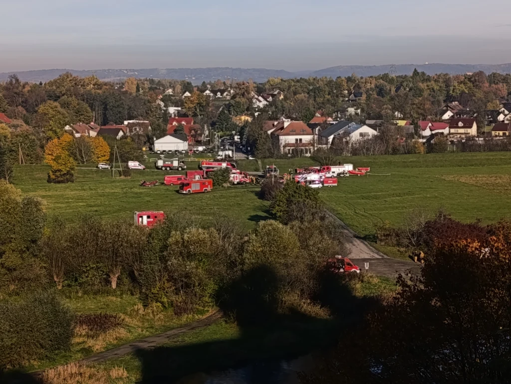 Kilka wozów strażackich i ratowniczych ustawionych na zielonym polu w pobliżu zabudowań mieszkalnych, otoczonych drzewami i niską roślinnością, w tle widoczne niskie domy oraz delikatnie zarysowane wzgórza.