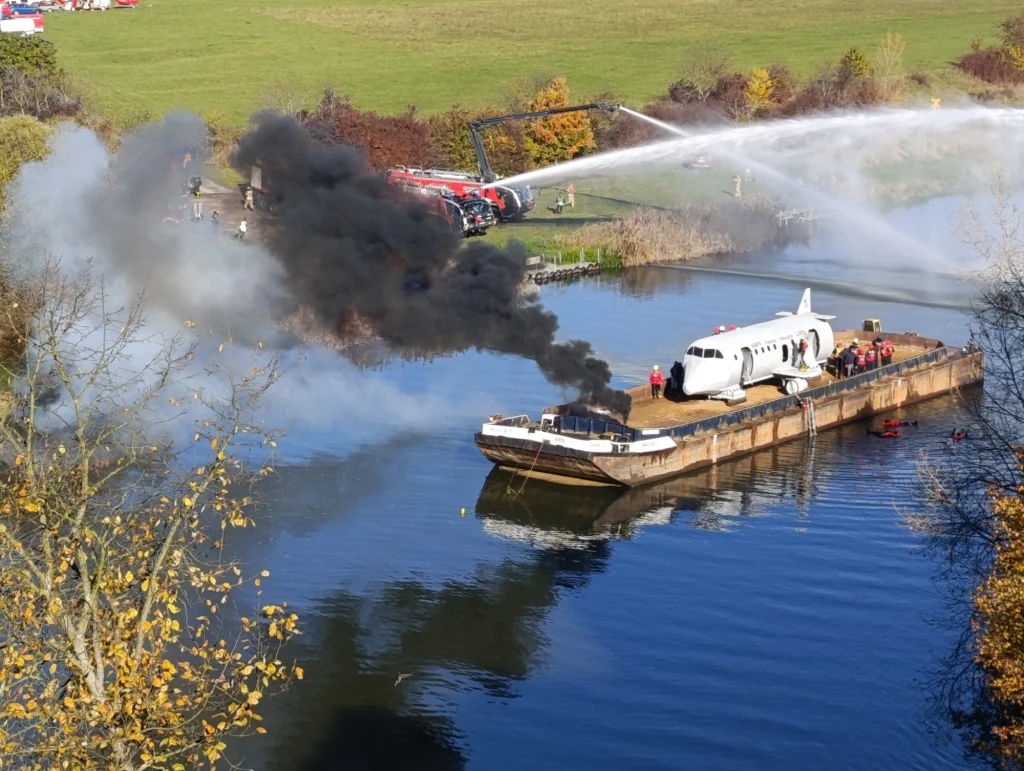 Ćwiartka samolotu umieszczona na barce na rzece, z której wydobywa się gęsty czarny dym. Strażacy na brzegu gaszą ogień wodą z armatki wodnej. W tle widać zieloną łąkę i pojazdy straży pożarnej.