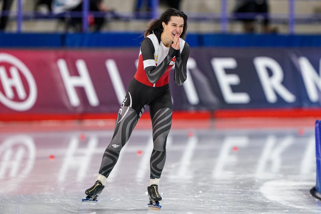 Łyżwiarka szybka w specjalnym stroju sportowym na torze lodowym, w tle rozmyte barierki i widownia, atmosfera sportowej rywalizacji.