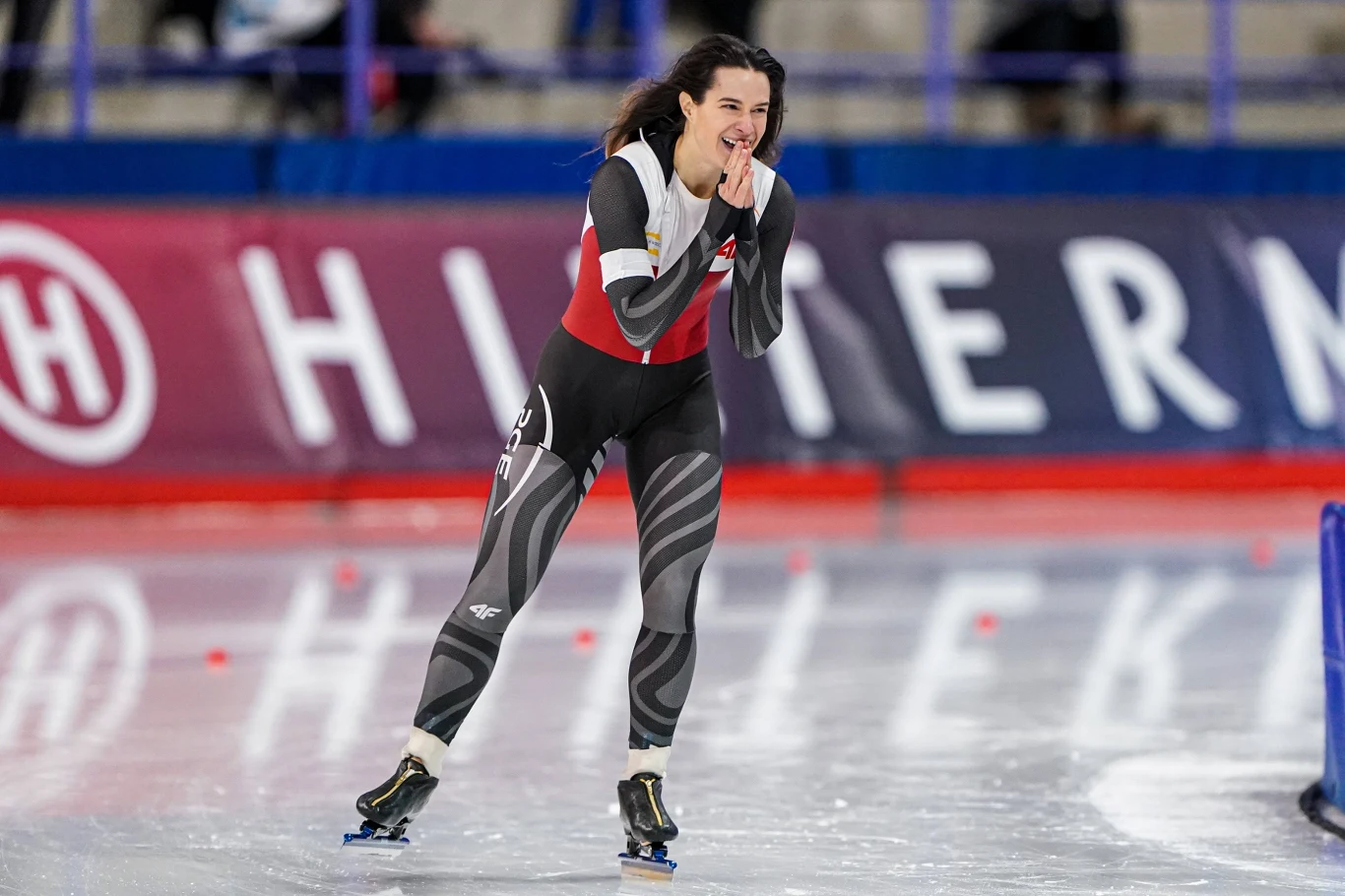 Łyżwiarka szybka w specjalnym stroju sportowym na torze lodowym, w tle rozmyte barierki i widownia, atmosfera sportowej rywalizacji.