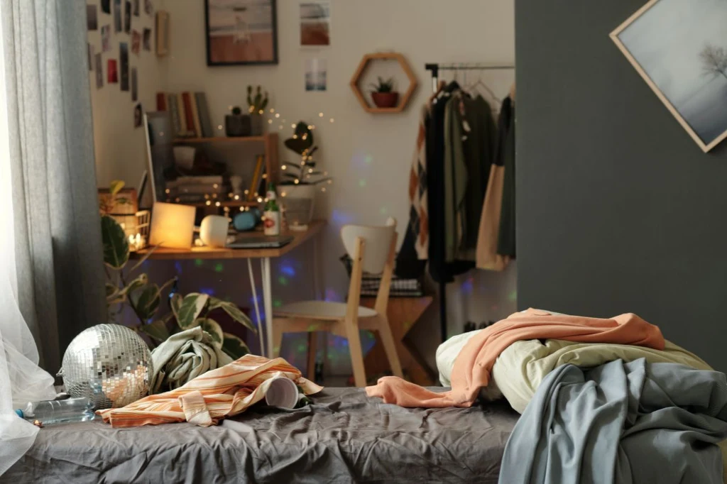 Przytulny pokój z nieposłanym łóżkiem, ubraniami, kulą dyskotekową, biurkiem z lampką i dekoracjami, w tle rośliny doniczkowe i ozdobne światełka, na ścianach wiszą obrazy i zdjęcia, a na stojaku wisi kilka ubrań.