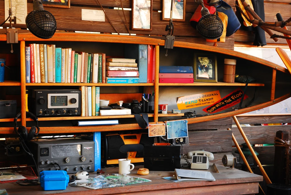 Drewniana półka w kształcie łódki wypełniona książkami, sprzętem elektronicznym, starym telefonem, radiem, zdjęciami oraz drobiazgami, które tworzą atmosferę retro pracowni lub gabinetu.