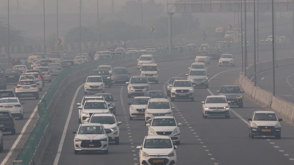 Gęsty ruch samochodowy na kilku pasmach autostrady, w tle widoczna mgła lub smog, słaba widoczność i szare niebo.