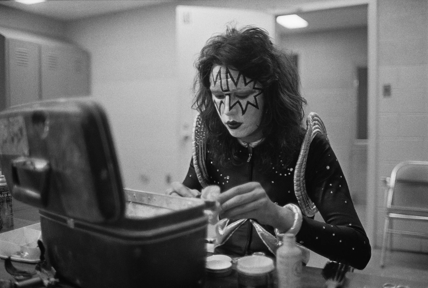 Ace Frehley w składzie Kiss w 1974 r. Osoba z efektownym makijażem scenicznym i kostiumem siedzi przy stole z otwartą walizką, nakładając kosmetyki lub nakładając makijaż. W tle widoczne są jasne szafki oraz puste krzesło, co sugeruje miejsce za kulisami lub garderobę.