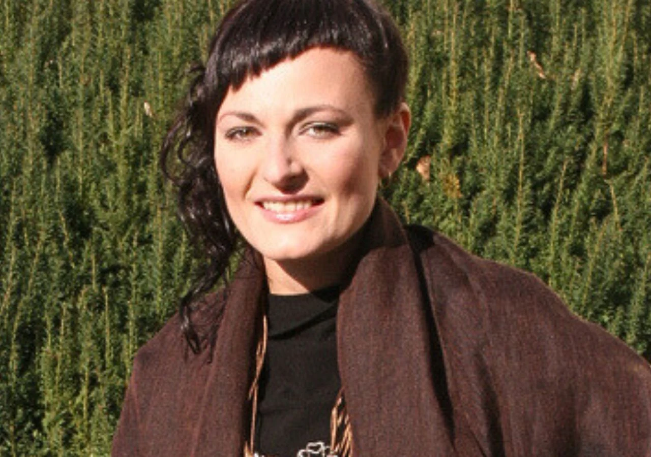 Miłka Skalska Kobieta z ciemnymi, krótko ściętymi włosami i nietuzinkową fryzurą uśmiecha się, stojąc na tle zielonych iglaków. Ubrana w ciemną kurtkę i szalik, spogląda w stronę aparatu.