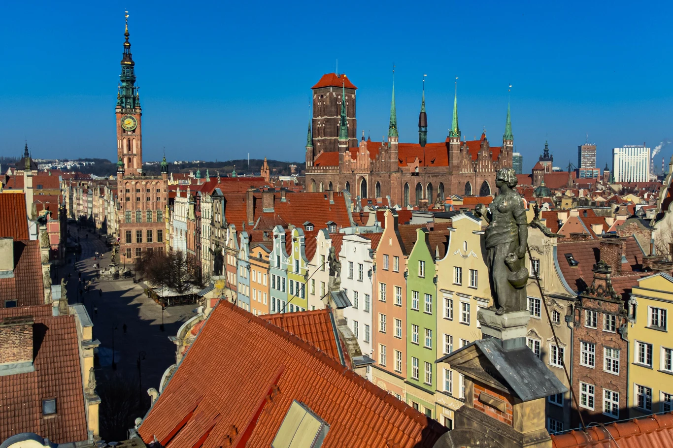 Urokliwe kawiarnie w Trójmieście Kolorowe kamienice starego miasta z charakterystycznymi czerwonymi dachami, monumentalny kościół Mariacki w tle oraz wieża ratusza, całość pod błękitnym niebem i z widoczną rzeźbą na pierwszym planie.