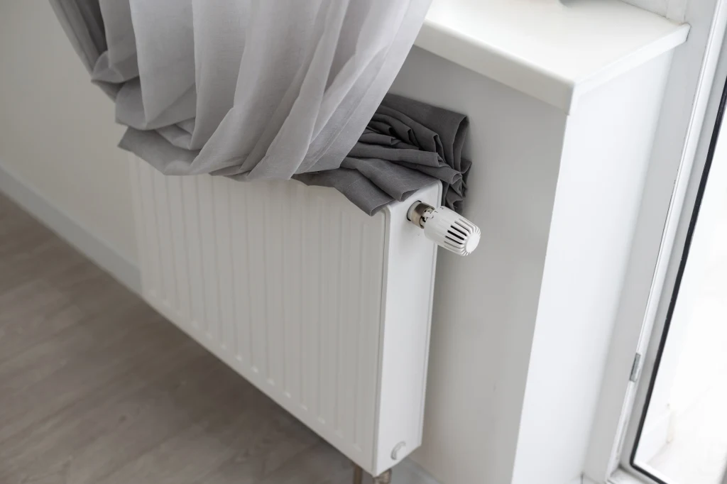 Biały grzejnik umieszczony pod parapetem, na górze grzejnika częściowo leży zwinięta zasłona oraz firana, obok znajduje się biała ściana i okno balkonowe.