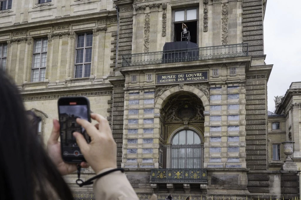 Fasada budynku muzeum Louvre, na pierwszym planie osoba trzymająca telefon i robiąca zdjęcie. Nad wejściem widoczny otwarty balkon oraz postać stojąca przy balustradzie.