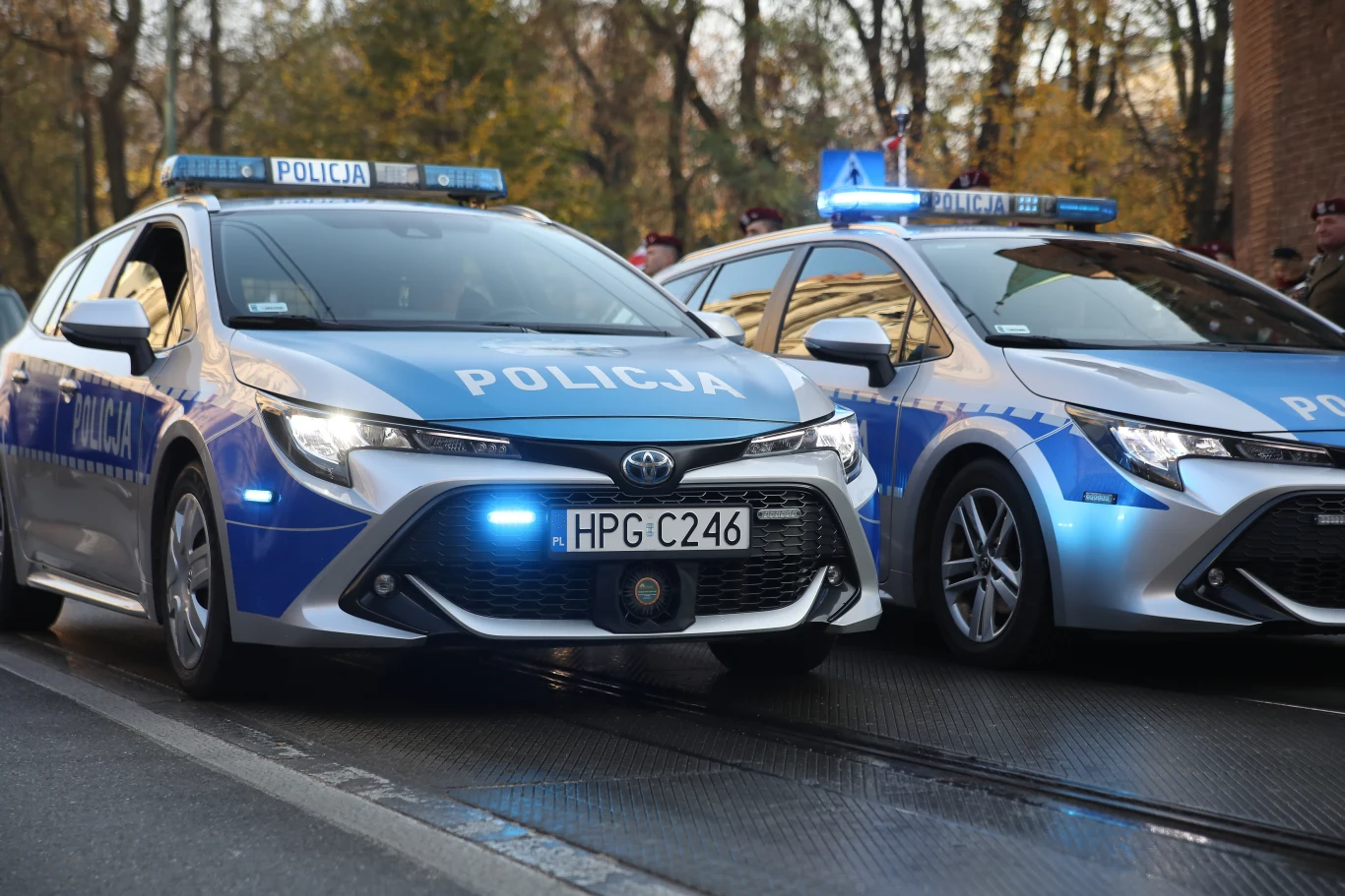 Śmierć 1,5-rocznej dziewczynki z Kołobrzegu. Sekcja nie wskazała przyczyny (zdj. ilustracyjne) Radiowozy policyjne marki Toyota z włączonymi światłami błyskowymi stoją na ulicy w otoczeniu drzew oraz skupiska ludzi w tle.