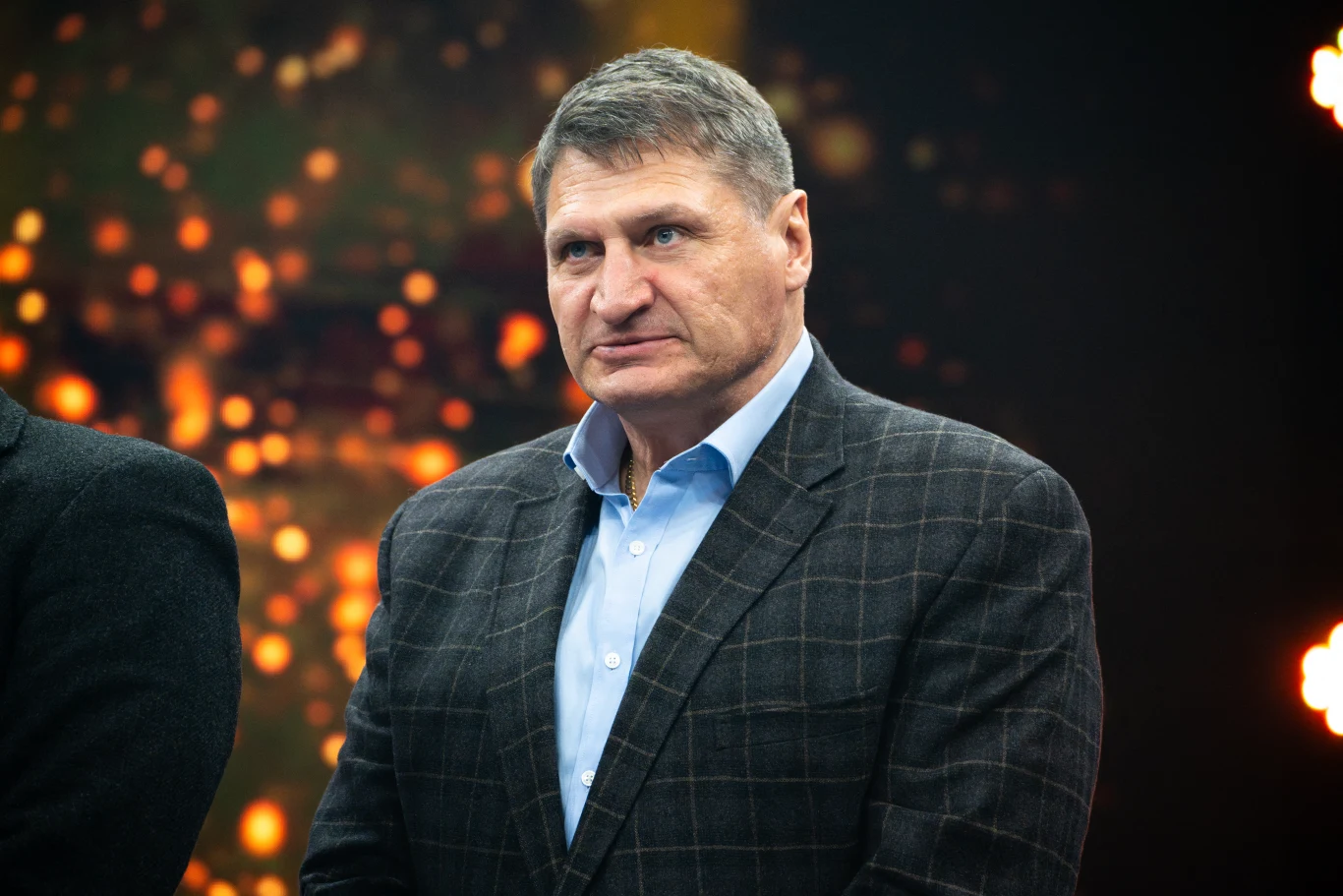 Mężczyzna w średnim wieku ubrany w elegancką marynarkę w kratę oraz jasnoniebieską koszulę, stoi na tle rozmytego, ciemnego tła z jasnymi punktami świetlnymi tworzącymi efekt dekoracyjny.